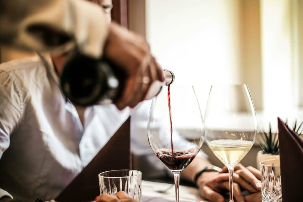 En person häller upp rödvin från en flaska i ett glas, med en annan person i bakgrunden och ett glas vitt vin på bordet.