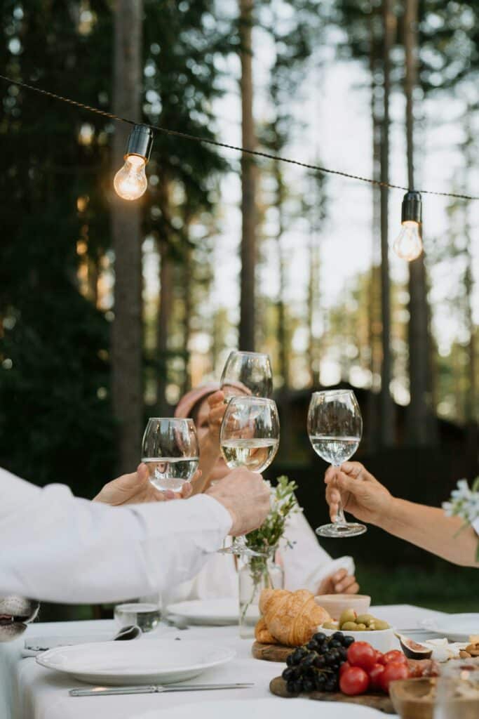 En grupp personer skålar med vinglas fyllda med vitt vin runt ett bord utomhus, omgivna av tallar och ljusslingor.